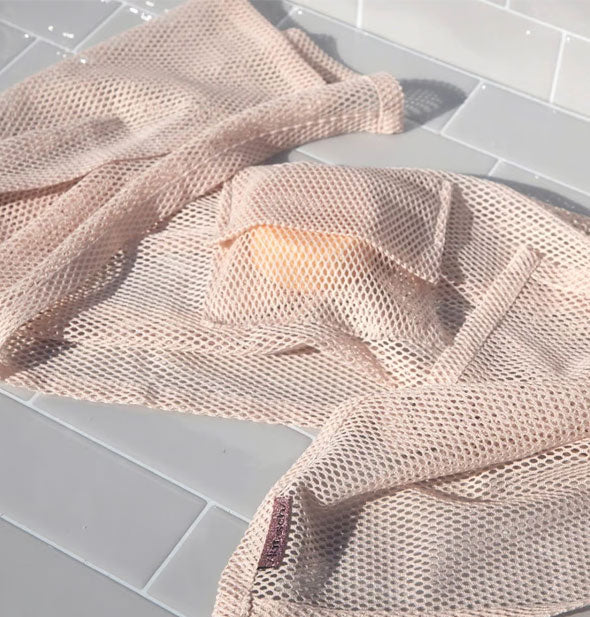 Light pink mesh washcloth with soap bar inside pocket lays on a white tiled surface