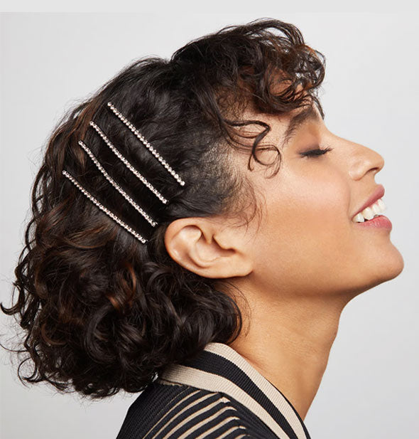 Profile of smiling model with eyes closed shows four rhinestone hair pins in a side-swept hairstyle