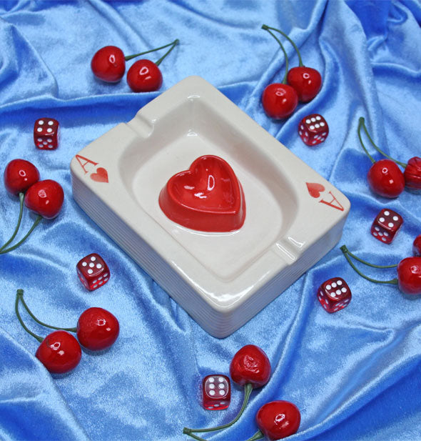 Ace of hearts card ashtray rests on shiny blue fabric scattered with red cherries and dice