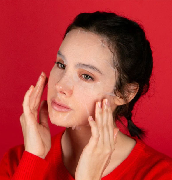 Model wearing red against a red backdrop smooths a sheet mask over face with fingertips
