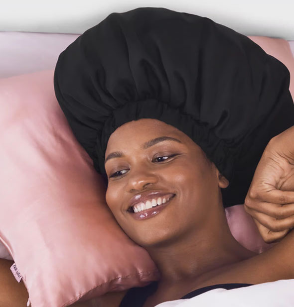Smiling model wearing a black XL Satin Bonnet rests on a pink satin pillow
