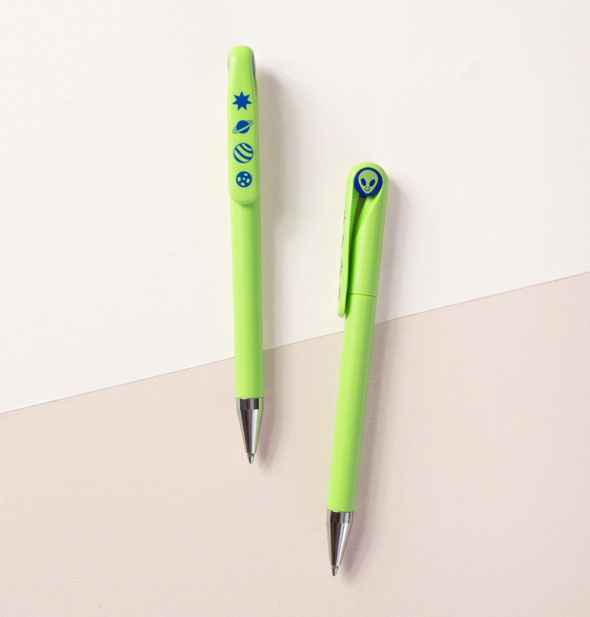 Two green alien pens on a two-tone neutral surface feature dark blue celestial and supernatural graphics