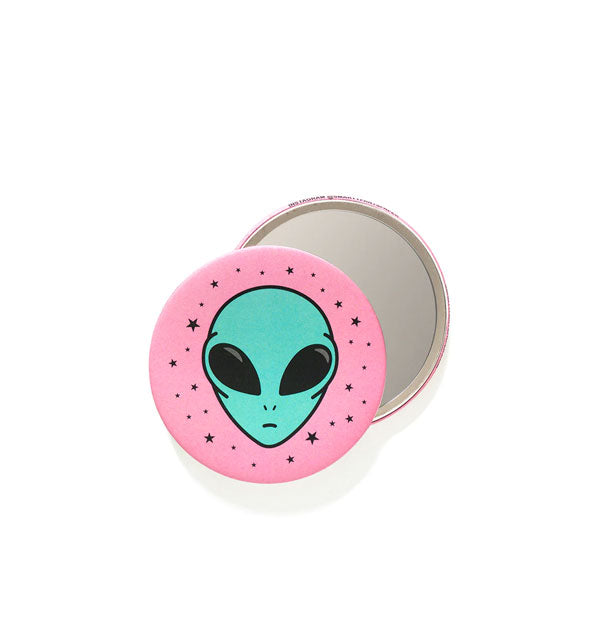Front and back views of a round pocket mirror featuring a teal alien head illustration surrounded by small stars on a pink background