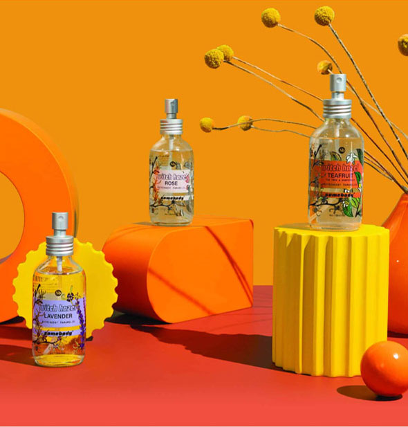 Collection of Lavender, Rose, and Teafruit Witch Hazel sprays on a bright orange and yellow backdrop staged with geometric shapes and flower sprigs