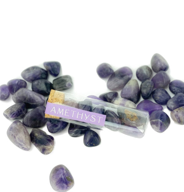 Clear vial of polished purple amethyst stones with cork cap and printed purple label rests on top of other loose amethyst stones