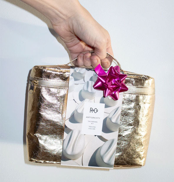 Model's hand holds a metallic gold R+Co Antigravity kit bag with metallic pink ribbon attached by its handle