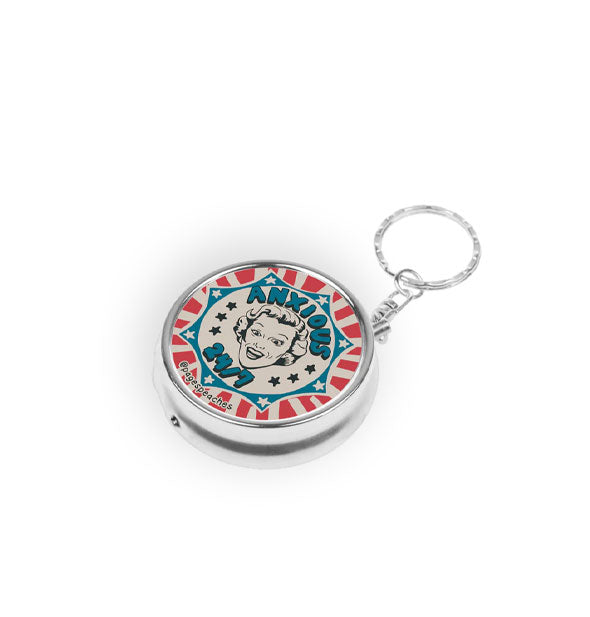 Round silver pill case on keychain features red, white, and blue artwork with a woman's surprised face in the center and the phrase, "Anxious 24/7" in blue lettering