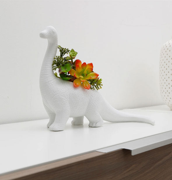 White porcelain long-necked dinosaur vase on office cabinet holds a small arrangement of flowers and succulents