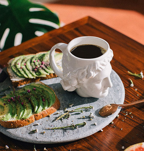 White Apollo head mug filled with black coffee on a wooden tabletop with stone tray and slices of avocado toast