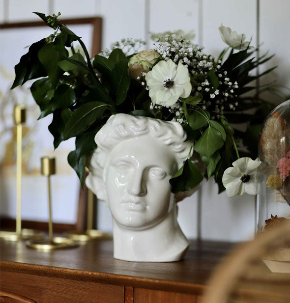White Apollo head vase on a wooden chest of drawers holds a bouquet of flowers and lots of greenery