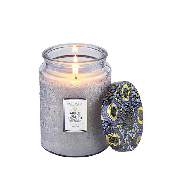 Lit Voluspa Apple Blue Clover candle in a light blue embossed glass jar with blue, gold, and black floral patterned tin lid removed and propped up alongside