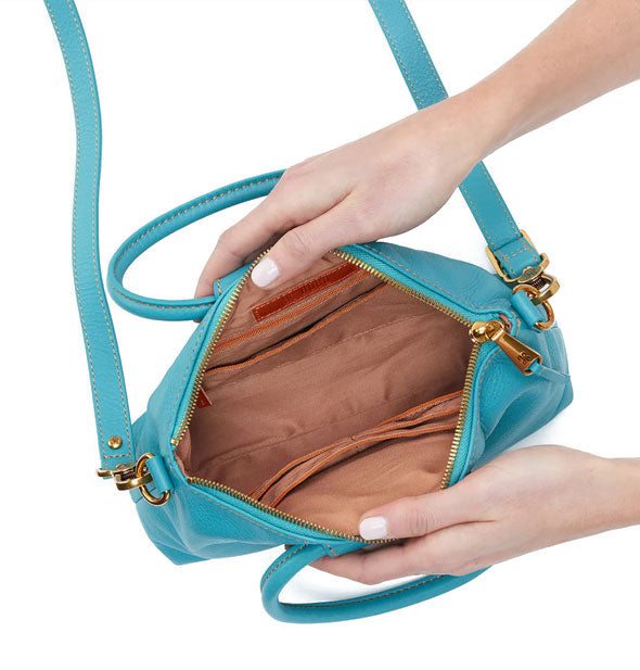 Model's hands hold open an aqua blue leather handbag to show brown interior with pocket storage