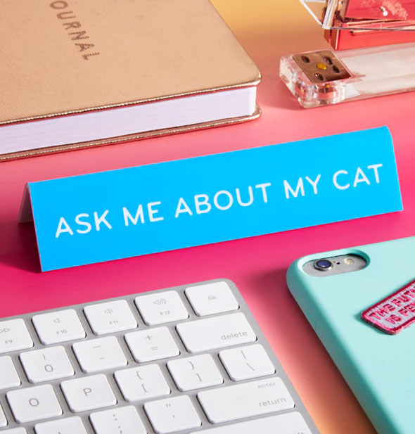 Blue "Ask Me About My Cat" desk sign is staged with a computer keyboard, smartphone, gold journal, and stapler on a pink surface
