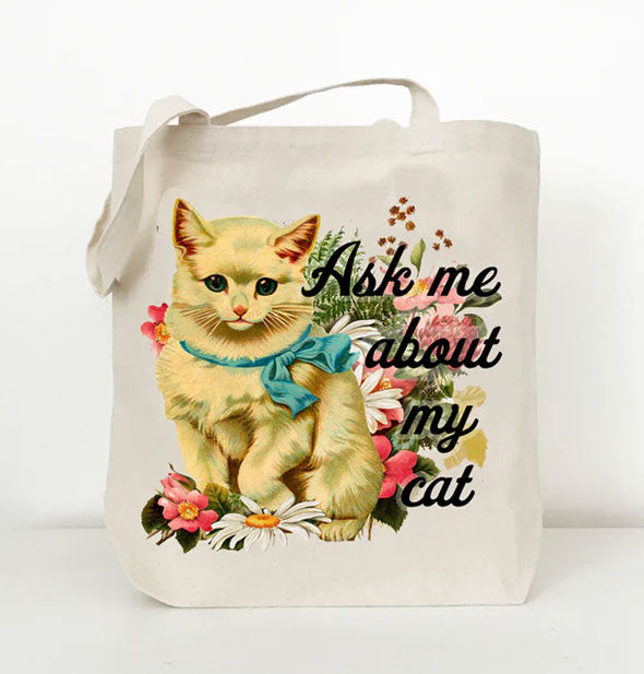 White cotton canvas tote bag features a colorful retro-style illustration of a yellow cat wearing a blue ribbon and surrounded by flowers next to the phrase, "Ask me about my cat" in black script