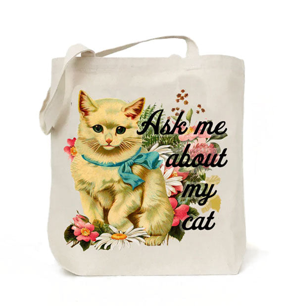 White cotton canvas tote bag features a colorful retro-style illustration of a yellow cat wearing a blue ribbon and surrounded by flowers next to the phrase, "Ask me about my cat" in black script