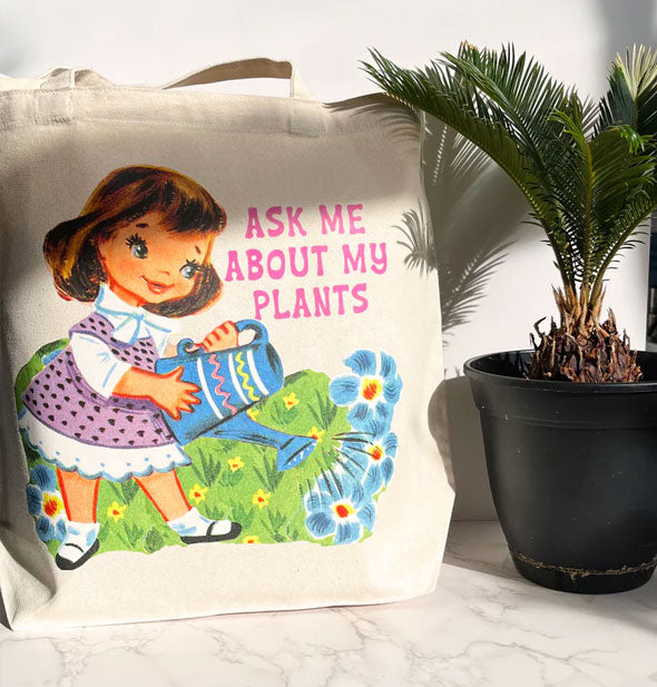 White canvas tote bag rests on a marble surface next to a potted palm tree and features colorful retro-style artwork of a little girl watering flowers next to the words, "Ask me about my plants" in pink lettering
