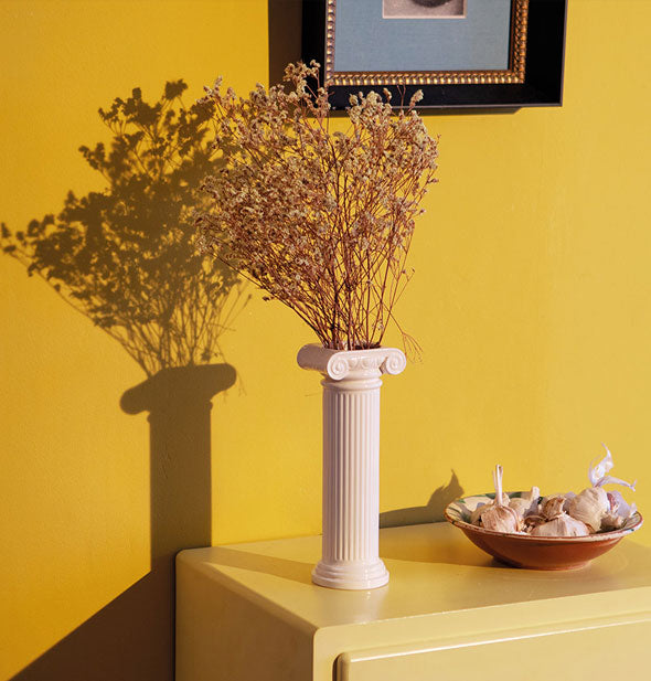 White column vase on top of a chest holds a bouquet of dried flowers against a bright yellow wall