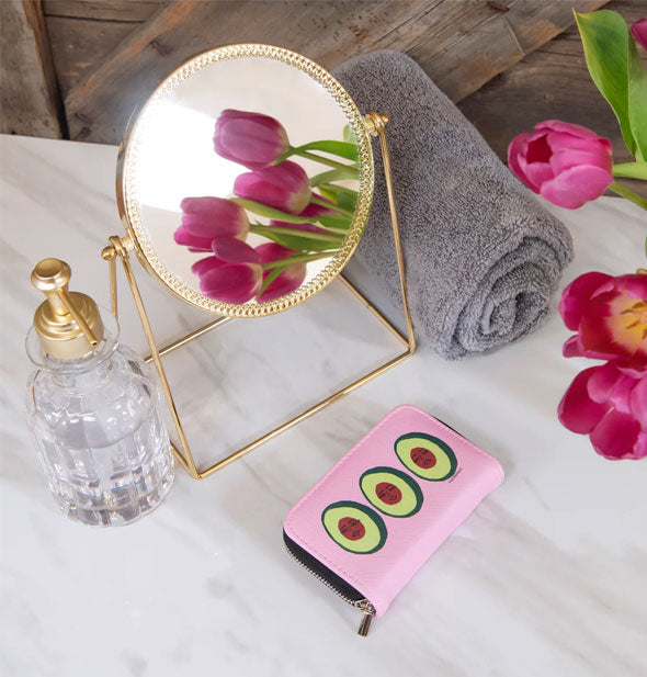 Avocadoze manicure kit pouch on a marble tabletop with mirror, crystal bottle, pink flowers, and rolled gray towel