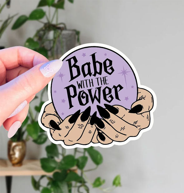 Model's hand holds a sticker depicting two hands with long, pointy, black fingernails holding a purple crystal ball that says, "Babe With the Power" surrounded by faint star accents