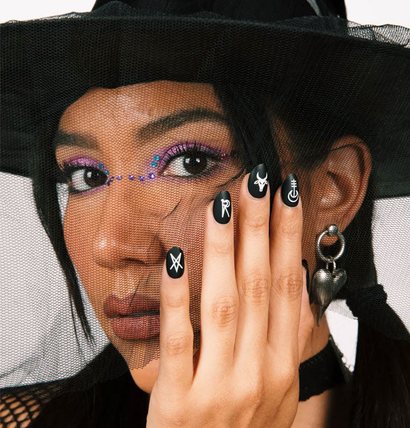 Model in witch hat with veil holds hand to face showing black and white rune design press-on nails