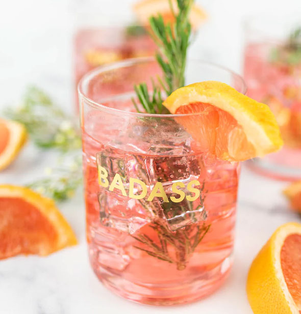 Badass rocks glass is filled with an iced pink beverage garnished with citrus and fresh herbs