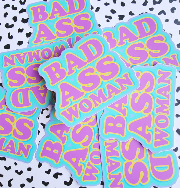 Pile of stickers that say, "Bad Ass Woman" in purple and yellow lettering on a teal background rest on a black and what speckled surface