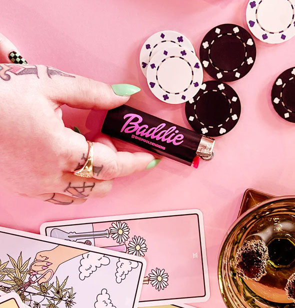 Model's hand touches a Baddie lighter on a pink surface with illustrated cards, a beverage, and black and white poker chips