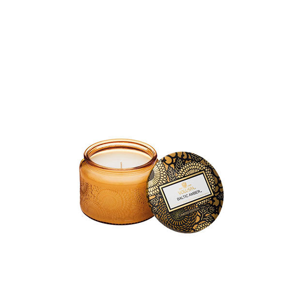 Golden embossed glass Baltic Amber Voluspa candle jar with black and gold metallic floral lid set to the side