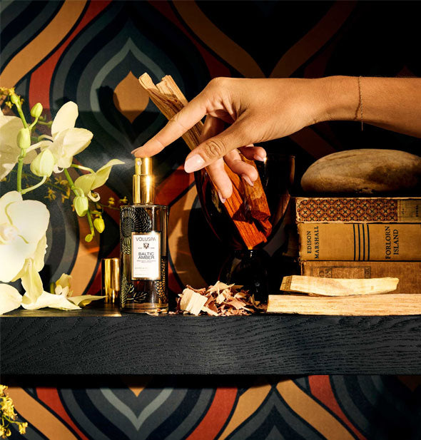 Model's finger presses down on the nozzle of a bottle of Voluspa Baltic Amber Room & Body Mist to dispense a fine spray against a backdrop of jewel-toned wallpaper, florals, and antique books