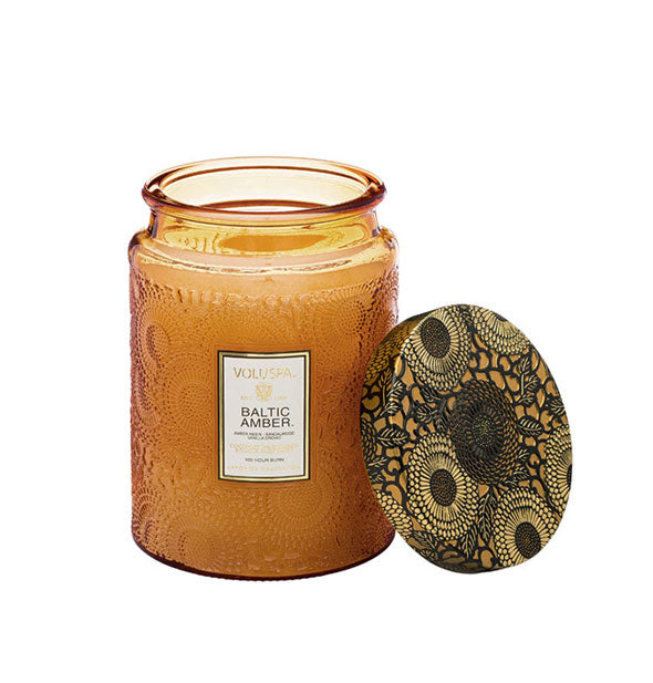 Golden embossed glass Baltic Amber Voluspa candle jar with black and gold metallic floral lid set to the side