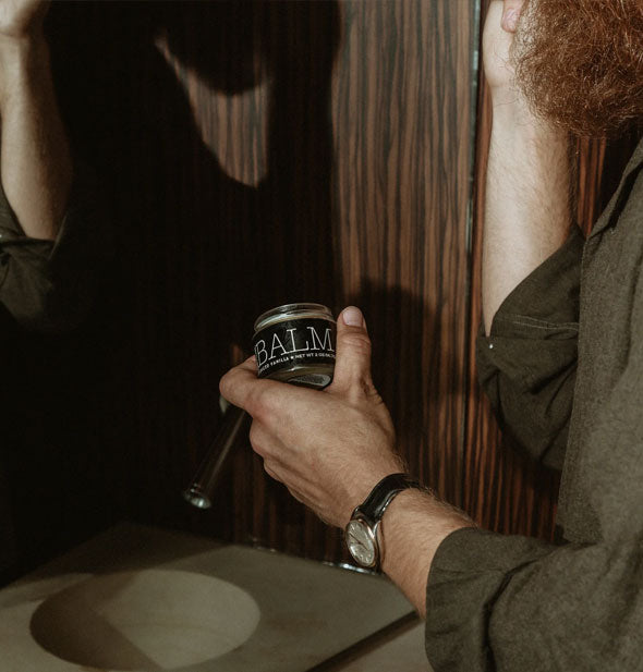 Model holding a jar of Balm for beards applies some product to facial hair while looking at a mirror