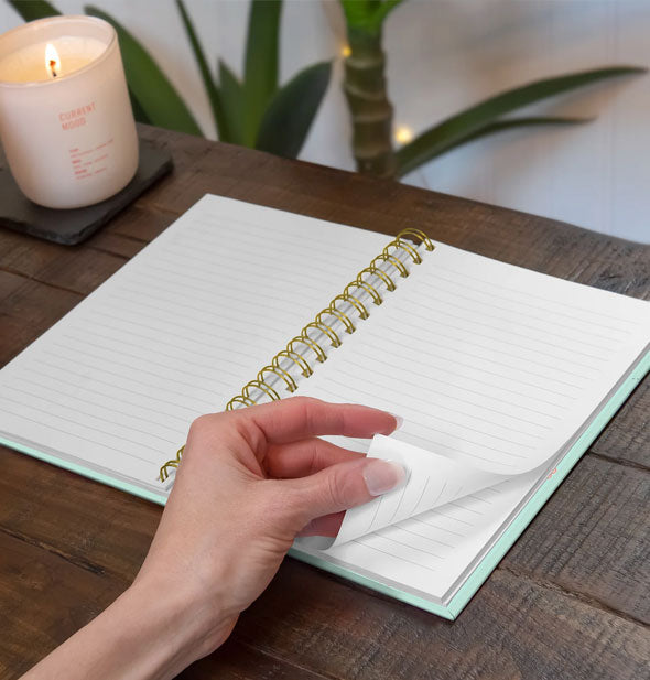 Model's hand turns the lined page of a spiral-bound notebook on a wooden desktop