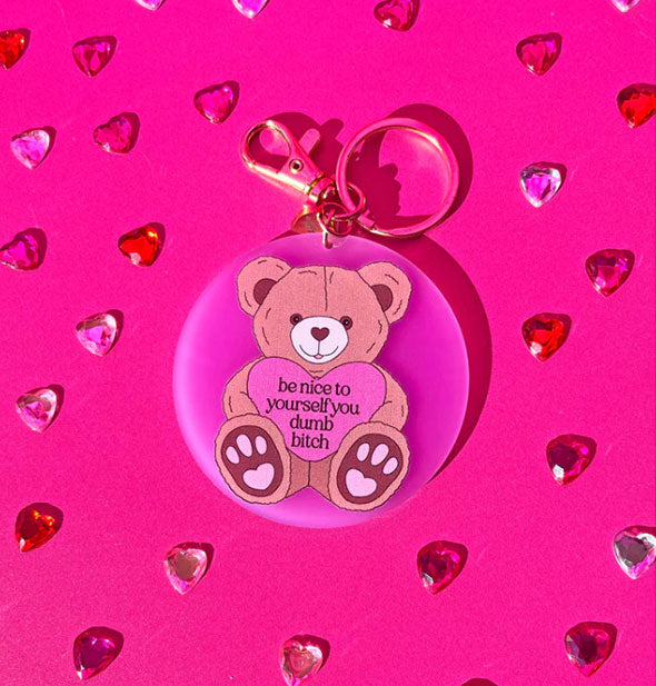 Round pink acrylic keychain on gold ring with clasp features illustration of a teddy bear holding a pink heart that says, "Be nice to yourself you dumb bitch" in black lettering. Surrounding it are small red, pink, and white crystal hearts