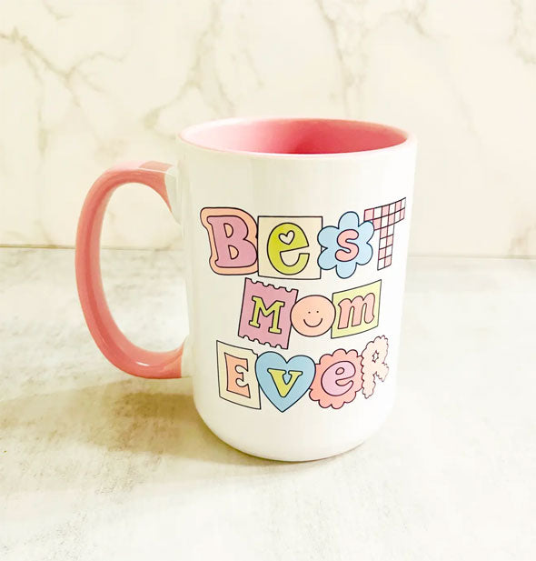 White ceramic coffee mug with pink handle and interior says, "Best mom ever" in alternating, fun letter styles in a pastel color palette