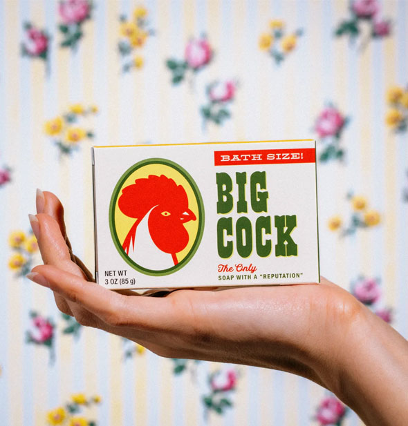 Model's hand holds a box of Big Cock bar soap up against a floral backdrop