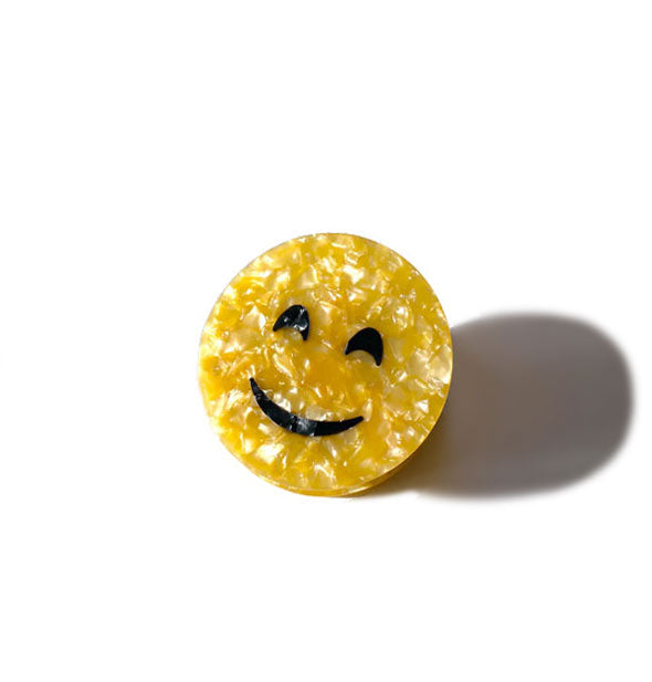 Round yellow quartz-effect hair clip with black smiley face design casts a harsh shadow on a light-colored surface