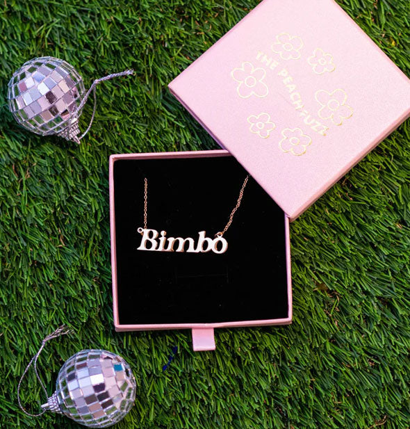 Gold "Bimbo" nameplate necklace rests on black felt in a purple slide-open box staged on green astroturf next to two mini disco balls