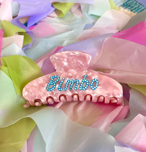 Pink quartz-effect hair claw clip with the word, "Bimbo" in blue rhinestones on its side rests on a pile of colorful ribbons