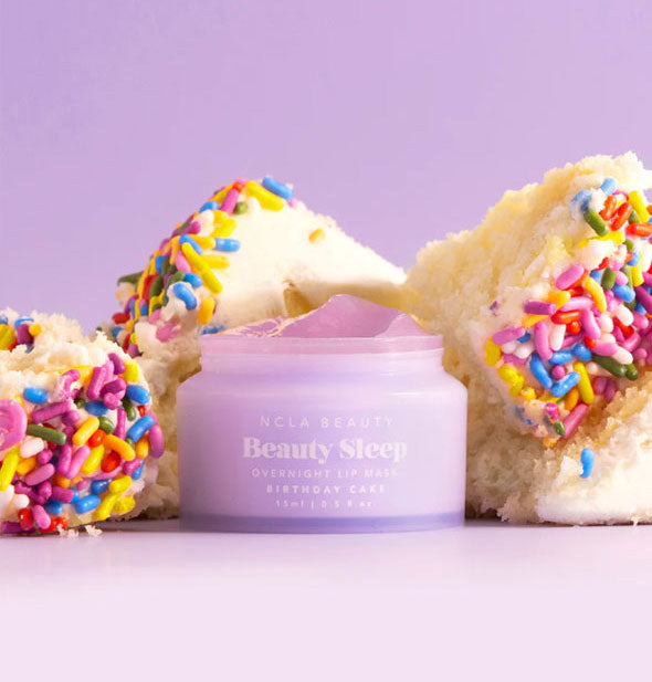 Opened purple pot of Birthday Cake NCLA Beauty brand Beauty Sleep Overnight Lip Mask staged with pieces of white cake with rainbow sprinkles