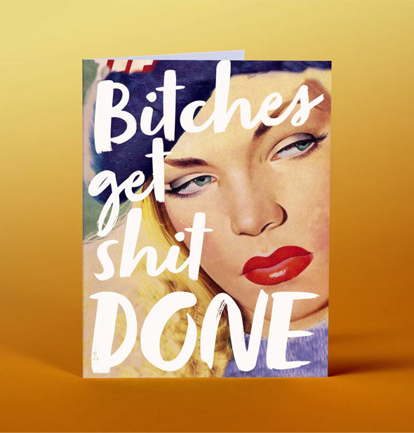 Greeting card on gold backdrop features closeup illustration of a retro-styled model's face alongside the words, "Bitches get shit done" in large white handwritten lettering
