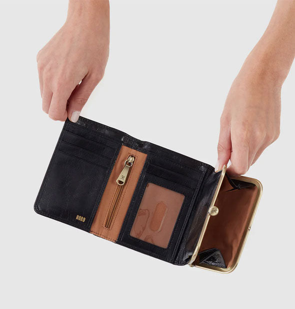 Model's hands hold open a black leather trifold wallet with brass hardware and brown lining