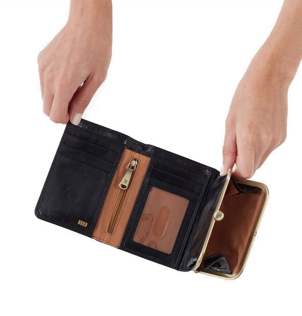 Model's hands hold open a black leather trifold wallet with brass hardware and brown lining