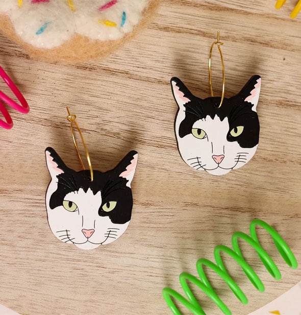 Black and white cat faces on gold earring hoops rest on a wooden surface with brightly colored springs and a plush toy