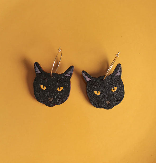 Pair of black cat faces with yellow eyes hand from gold earring hoops on a yellow backdrop