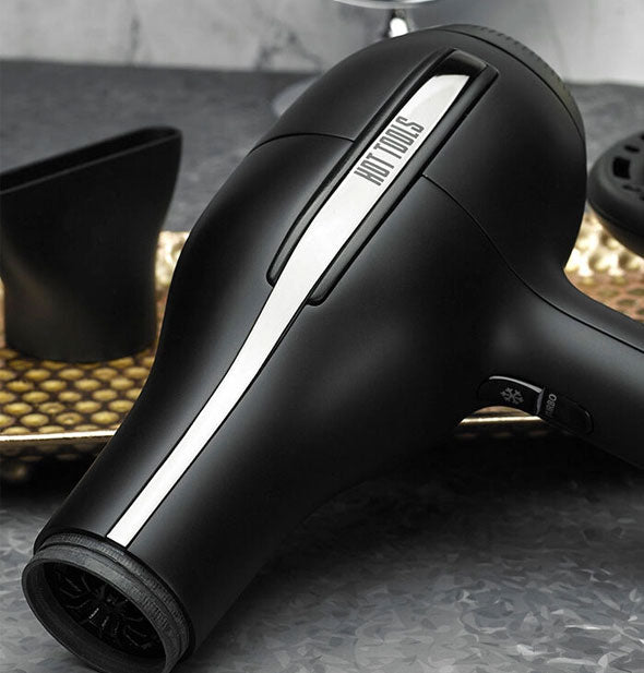 Hot Tools hair dryer rests on a vanity with diffuser and concentrator in the background on a gold tray