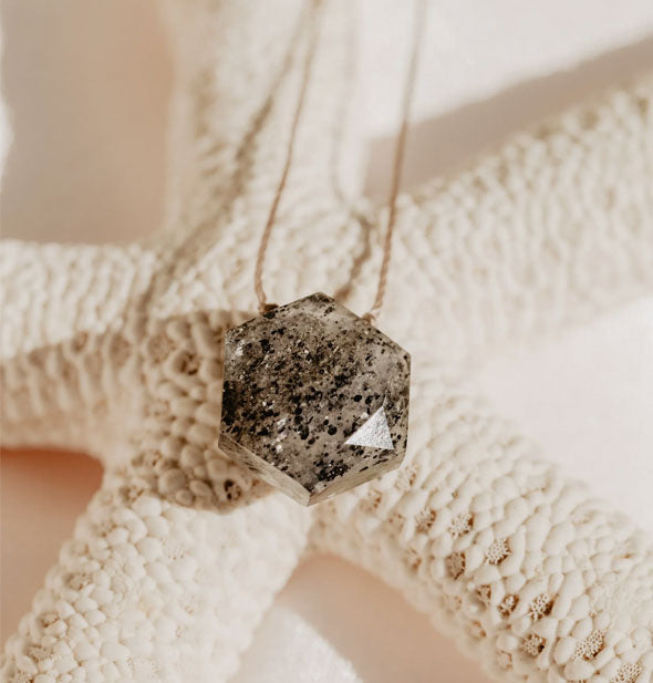 A speckled black sunstone hexagonal necklace on gold cord rests on a white starfish