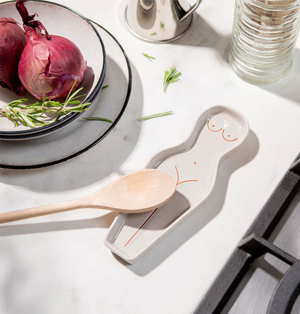 White ceramic spoon rest supports a wooden spoon next to a gas range on a marble countertop alongside a bowl of red onions and fresh herbs