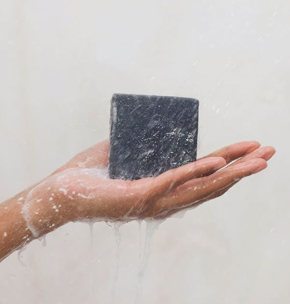 Model's hand holds a lathered bar of charcoal soap under a stream of shower water