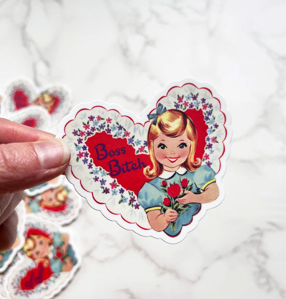 Model's hand holds a heart-shaped sticker featuring vintage-style artwork of a little girl holding red flowers against a floral border next to the words, "Boss Bitch"