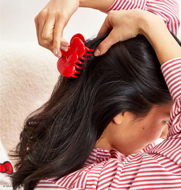 Model wearing a red and white striped shirt places a bright red bow-shaped claw clip into a partially-swept back hairstyle